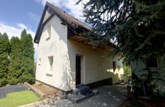 Záhradná chata so svetlou fasádou v obklopení zelene a ihličnatých stromov